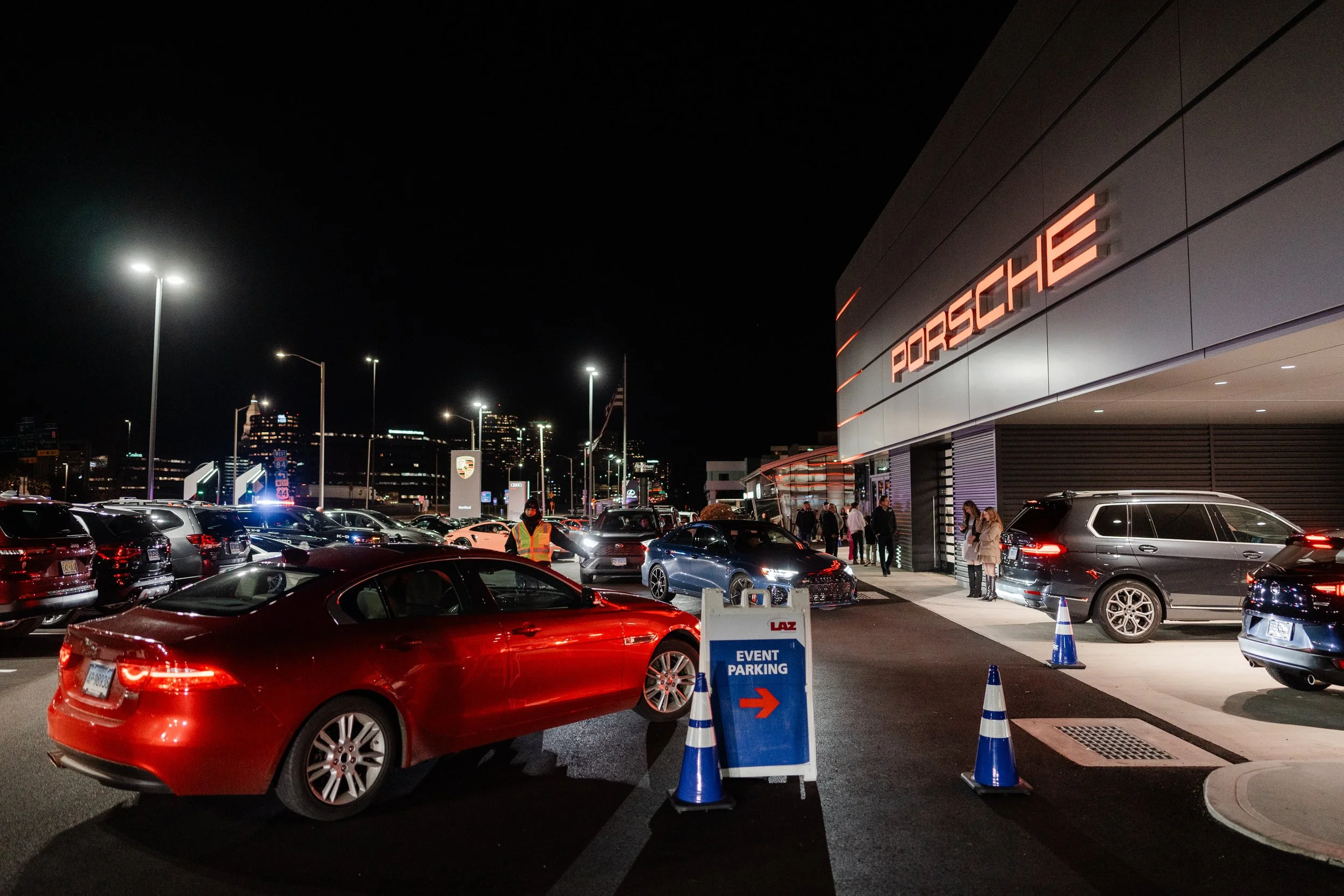 Nighttime scene outside Porsche dealership with multiple cars, a parking sign indicating event parking, and people walking or standing near the entrance.