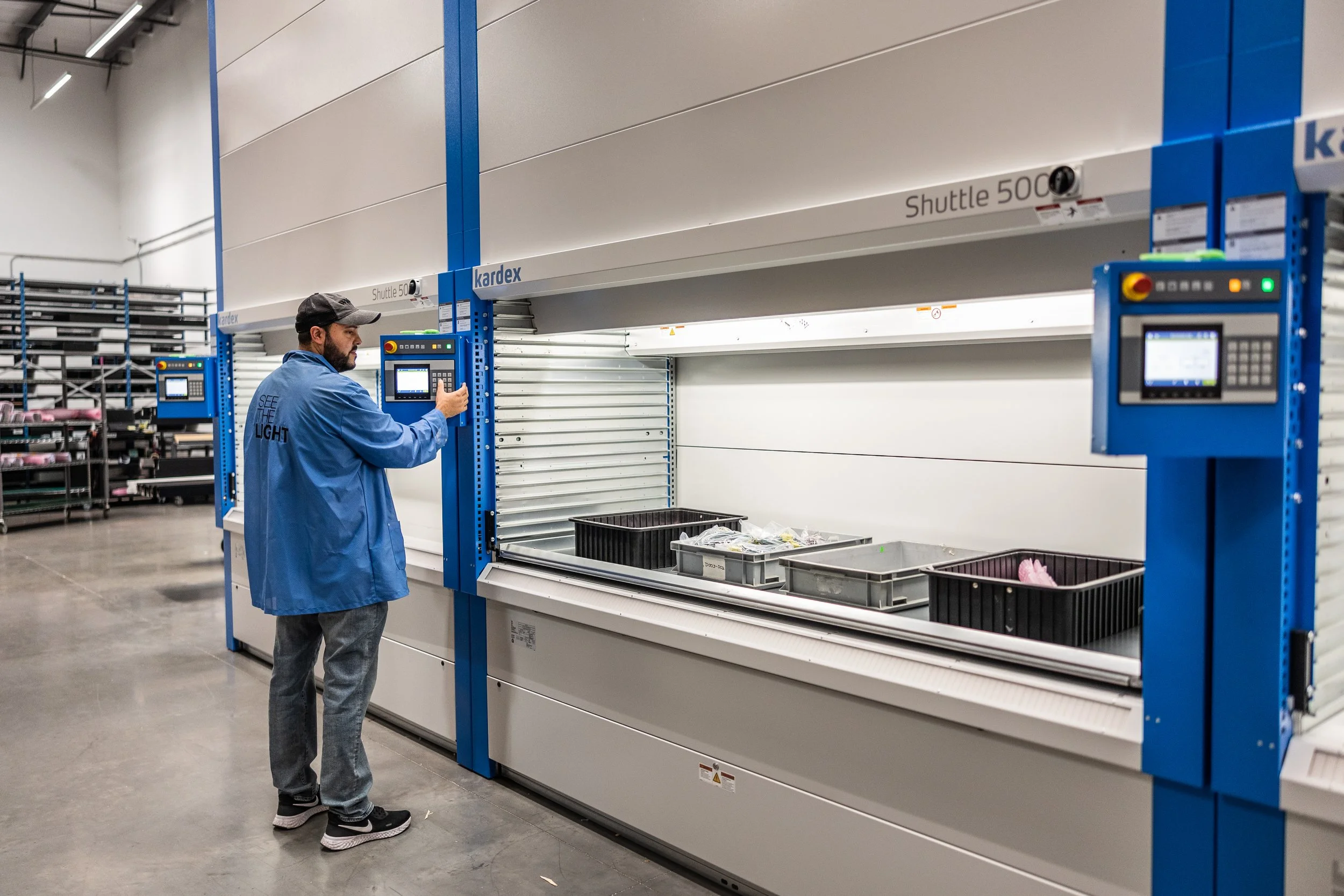 A worker wearing a blue jacket and gray pants is operating a large automated shelving or storage system in an industrial or warehouse setting. The system is labeled 'Shuttle 5000' and has a control panel that the worker is adjusting.