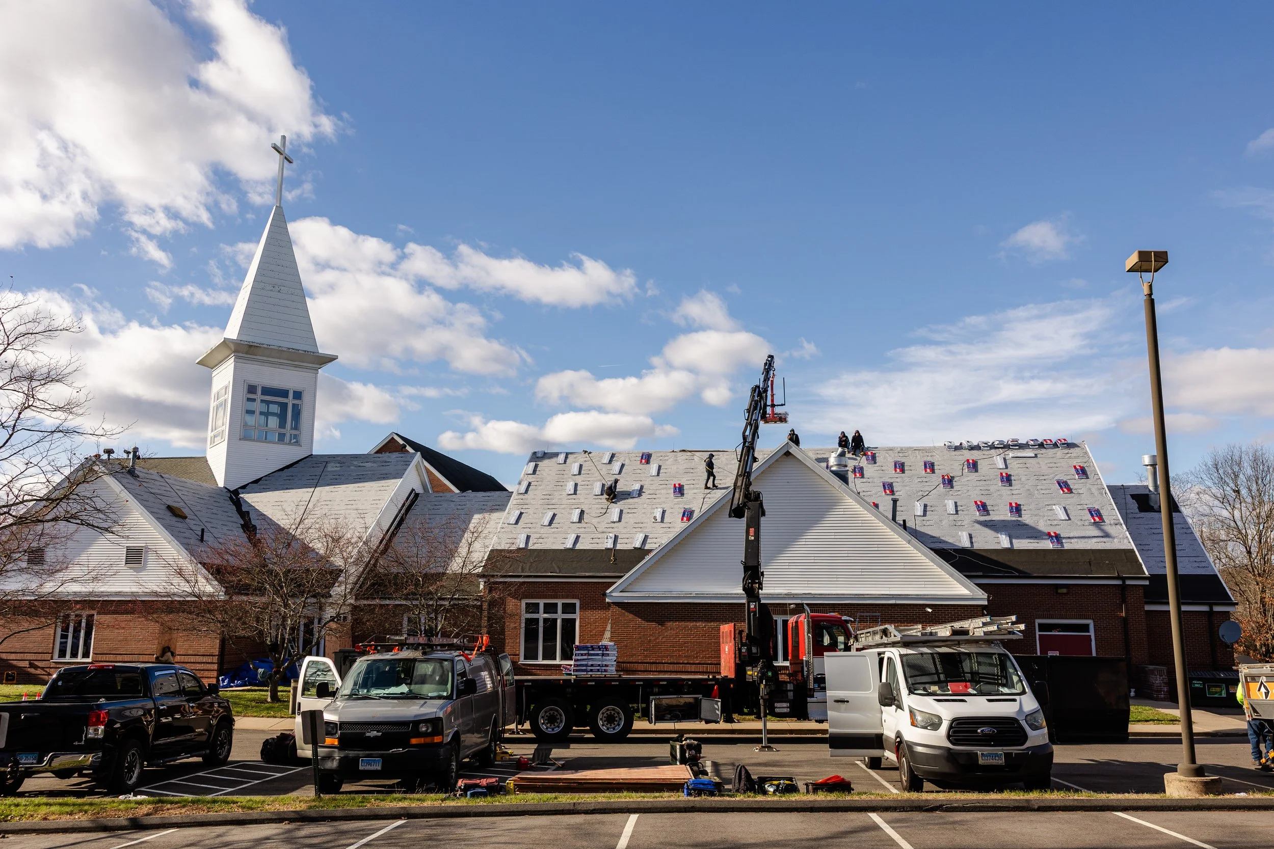A church with a steeple and a large roof under maintenance, with workers on the roof, trucks, and construction equipment parked outside on a sunny day.