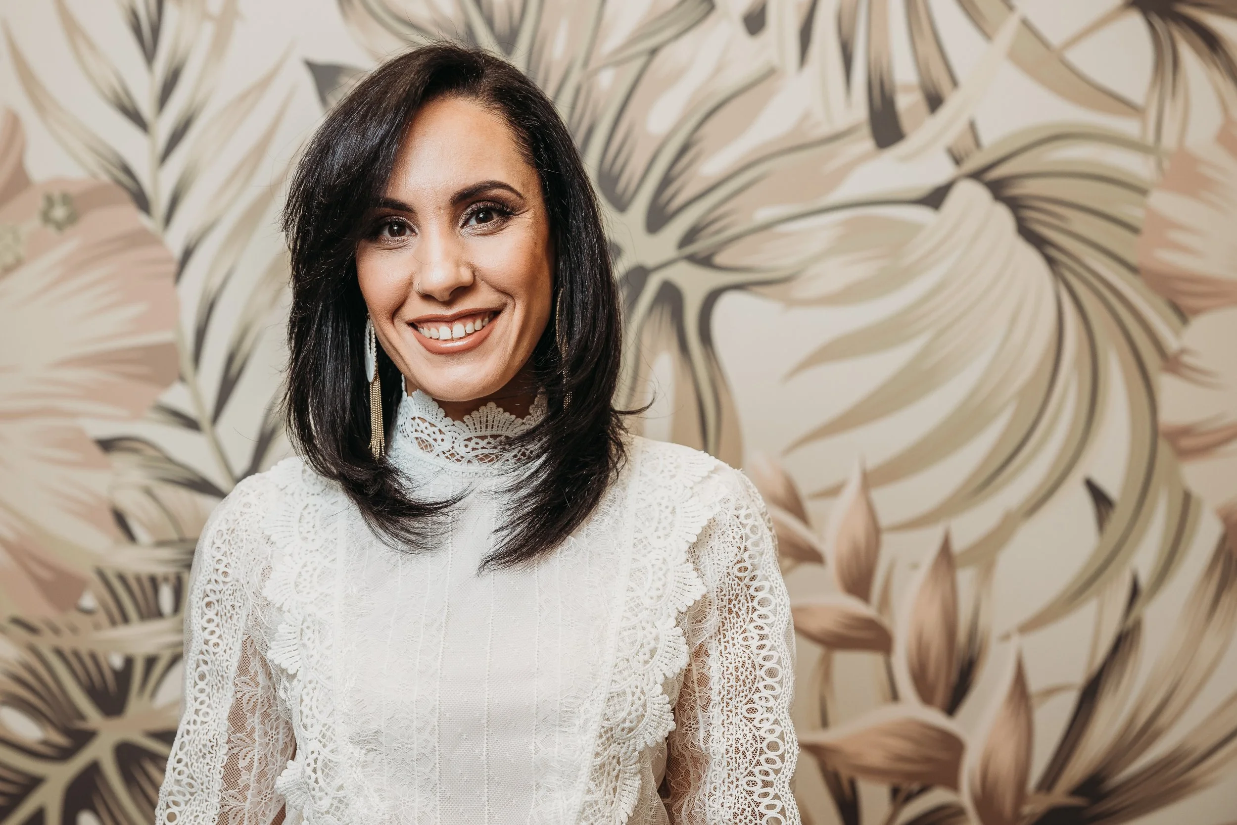 A woman with dark hair and earrings smiling in front of a beige and brown floral patterned wall, wearing a white lace high-neck dress.