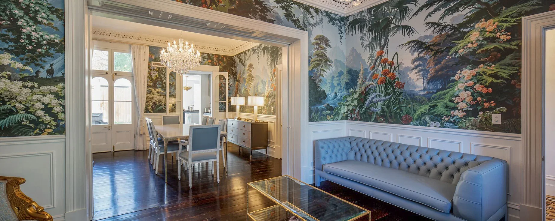 Elegant living room with vibrant jungle and mountain mural wallpaper, white wainscoting, tufted blue sofa, glass coffee table, and a doorway leading to a dining area with a chandelier and dark wood flooring.