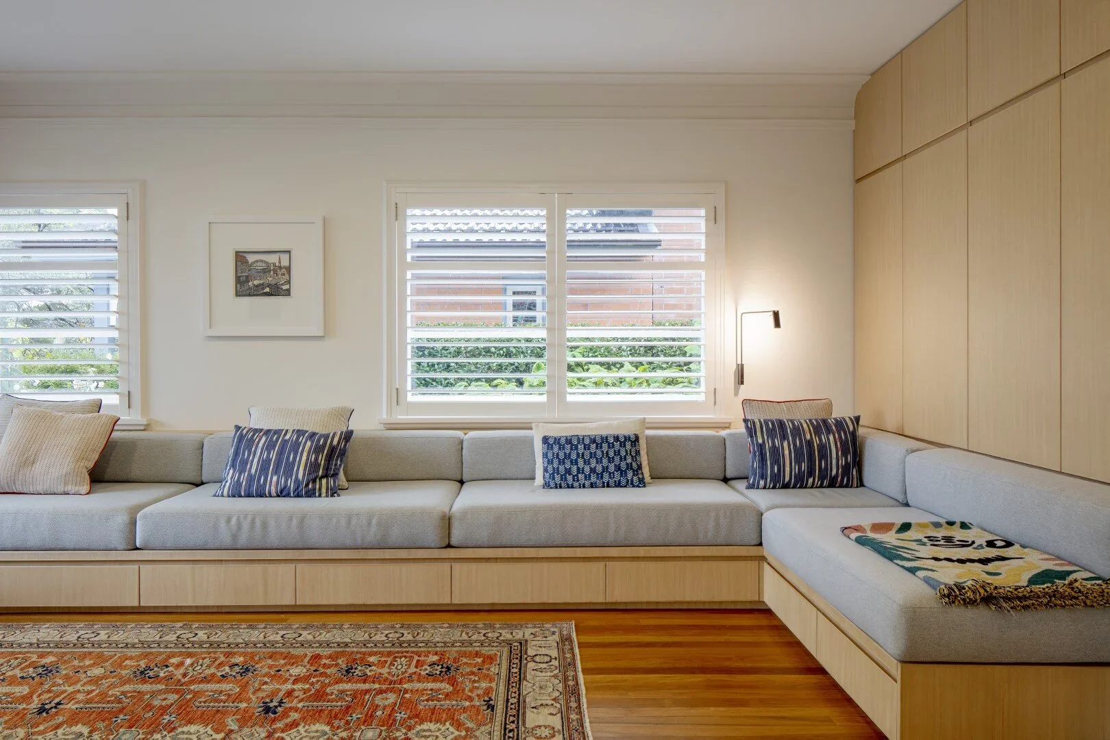 Living room with large L-shaped sofa, colorful pillows, wooden built-in storage, windows with white shutters, patterned area rug, and a wall-mounted reading lamp.