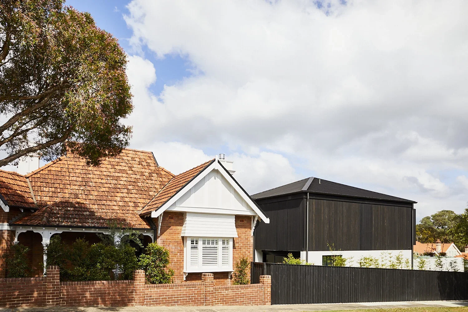 A residential neighborhood with traditional brick houses, one with a red tiled roof and white window shutters, and a modern black building behind a black fence, under a partly cloudy sky.