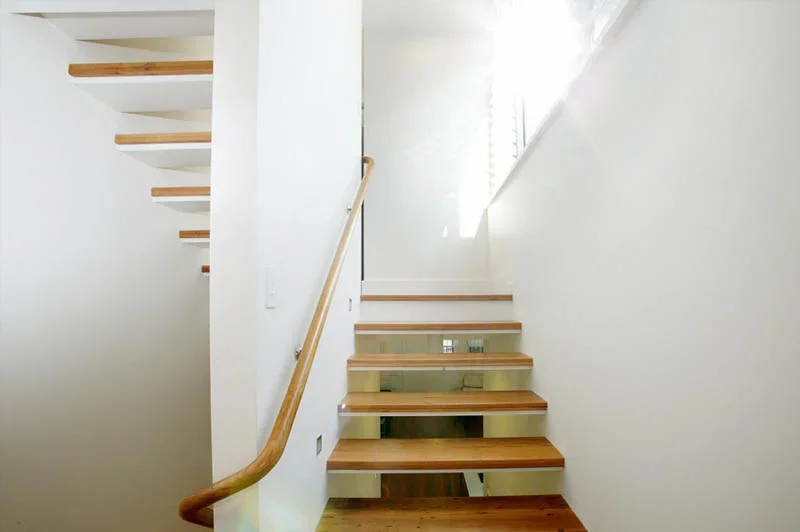 Interior view of a staircase with wooden steps and a light wood handrail, leading up to a second floor with a window allowing natural light to illuminate the space.