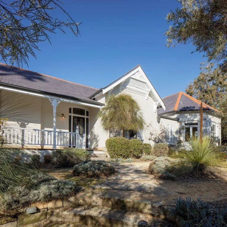 A charming white house with a front porch, surrounded by desert landscaping including bushes and trees, under a clear blue sky.
