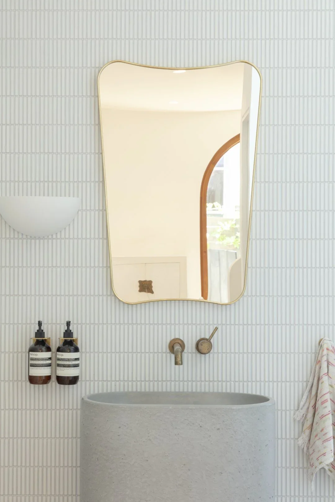 Minimalist bathroom with a curved stone sink, a large irregular-shaped mirror, wall-mounted faucet, bottles of soap, a towel on a hook, and a window in the reflection.