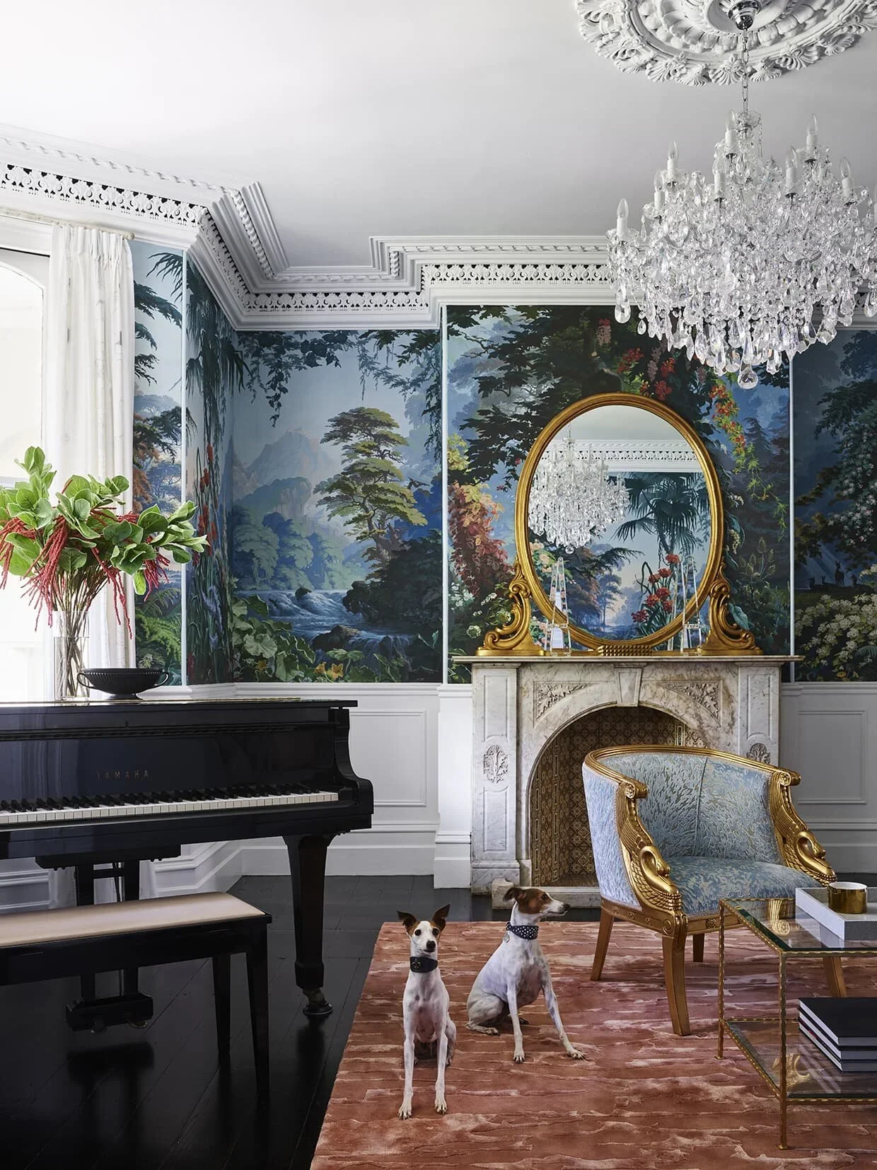 Elegant living room with ornate ceiling molding, large chandelier, mural of nature scene behind white marble fireplace with golden framed mirror, vintage blue armchair with gold accents, black Yamaha piano with a potted plant on top, two dogs sitting on a reddish-brown rug, and a glass coffee table with stacked books.