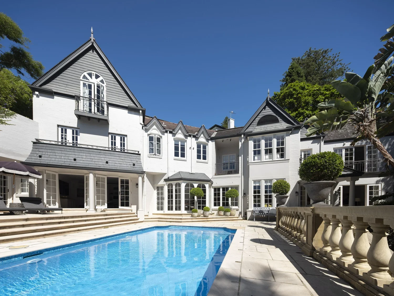 A large white house with multiple windows and balconies, a swimming pool in the backyard, and decorative plants and pots, under a clear blue sky.