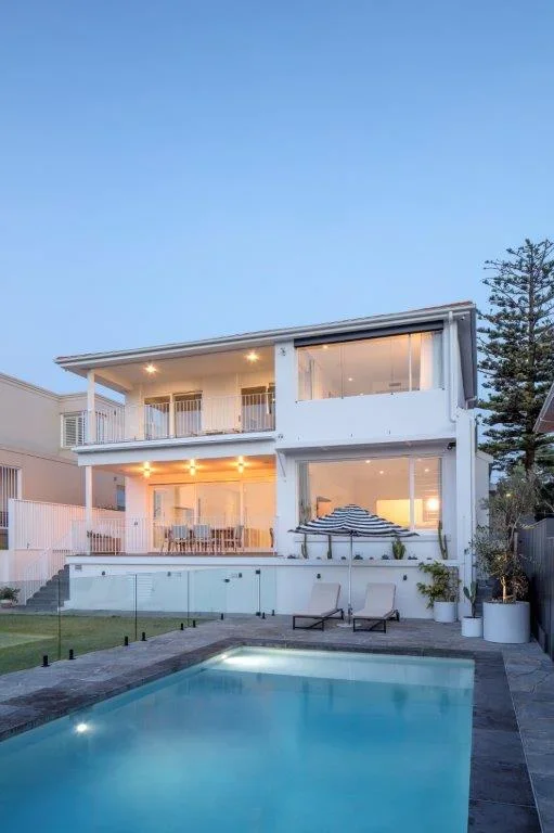 Modern white two-story house with large windows, balcony, patio, and a swimming pool in the backyard during twilight.