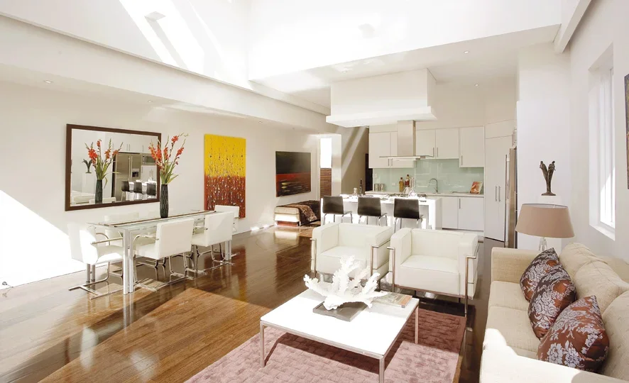 Modern open-concept living room and kitchen with white walls, wooden flooring, white furniture, and colorful artwork and décor.