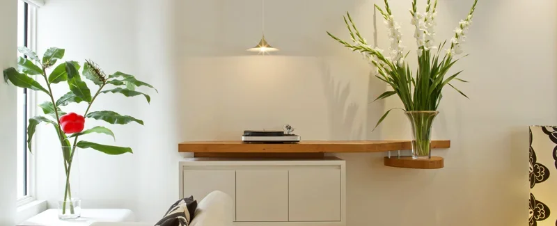 Interior living space with large green and red-leafed plant next to window, a wooden shelf with a tall glass vase of white flowers, and a white cabinet.