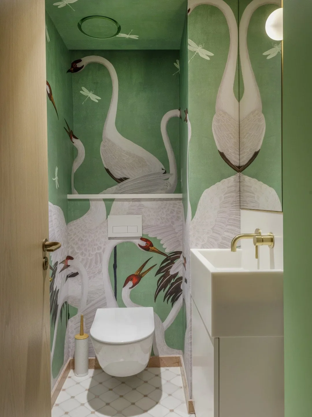 Decorative bathroom with green walls featuring large white cranes and dragonflies mural, white toilet, small white sink, gold faucet, and bathroom accessories.
