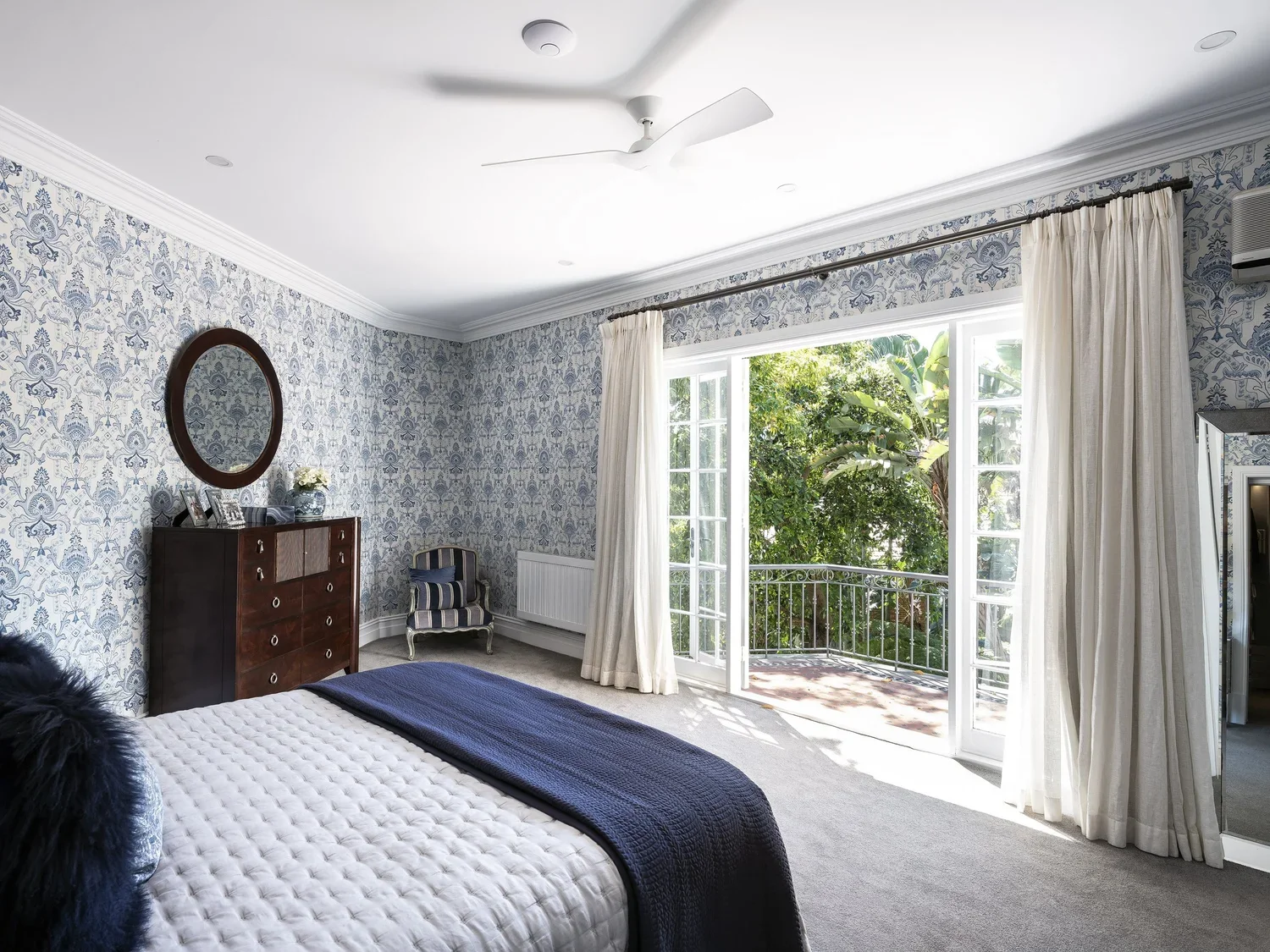 Bright bedroom with patterned blue wallpaper, large open window with beige curtains, and a balcony with greenery outside. Contains a bed with a white quilt and navy blanket, a wooden dresser with framed photos and a flower vase, a striped armchair, a