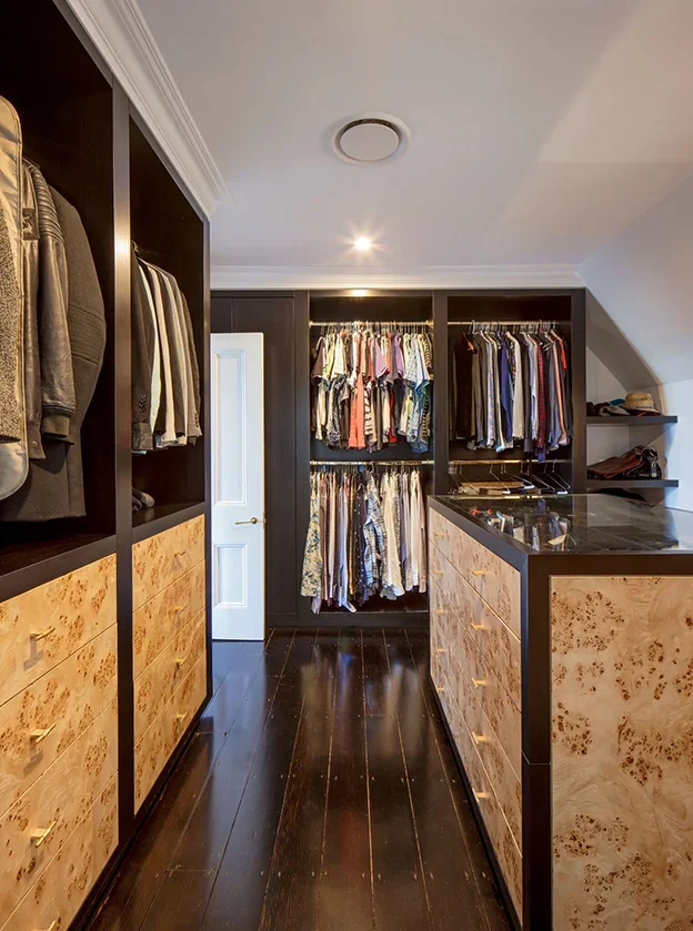 Walk-in closet with black shelving and drawers filled with clothing, including shirts and jackets, and a center island with marble top, in a room with dark wood flooring, crown molding, and a white door.
