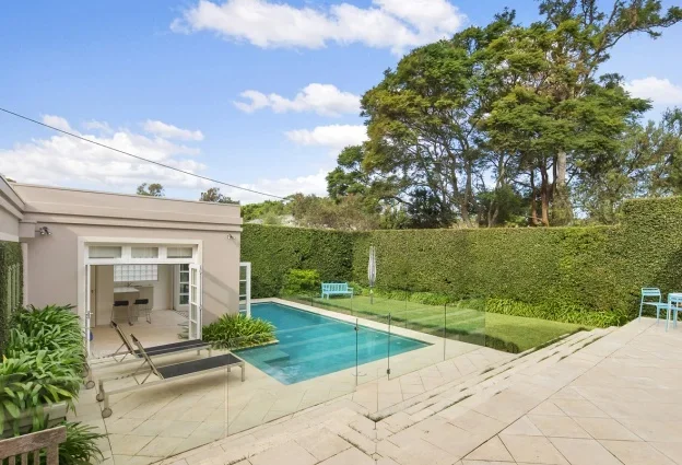 Backyard with a small pool, outdoor seating, a pergola, and a tall hedge with trees in the background.