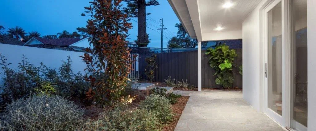 Modern backyard patio with plants, outdoor lighting, and a white exterior wall, taken during the evening.