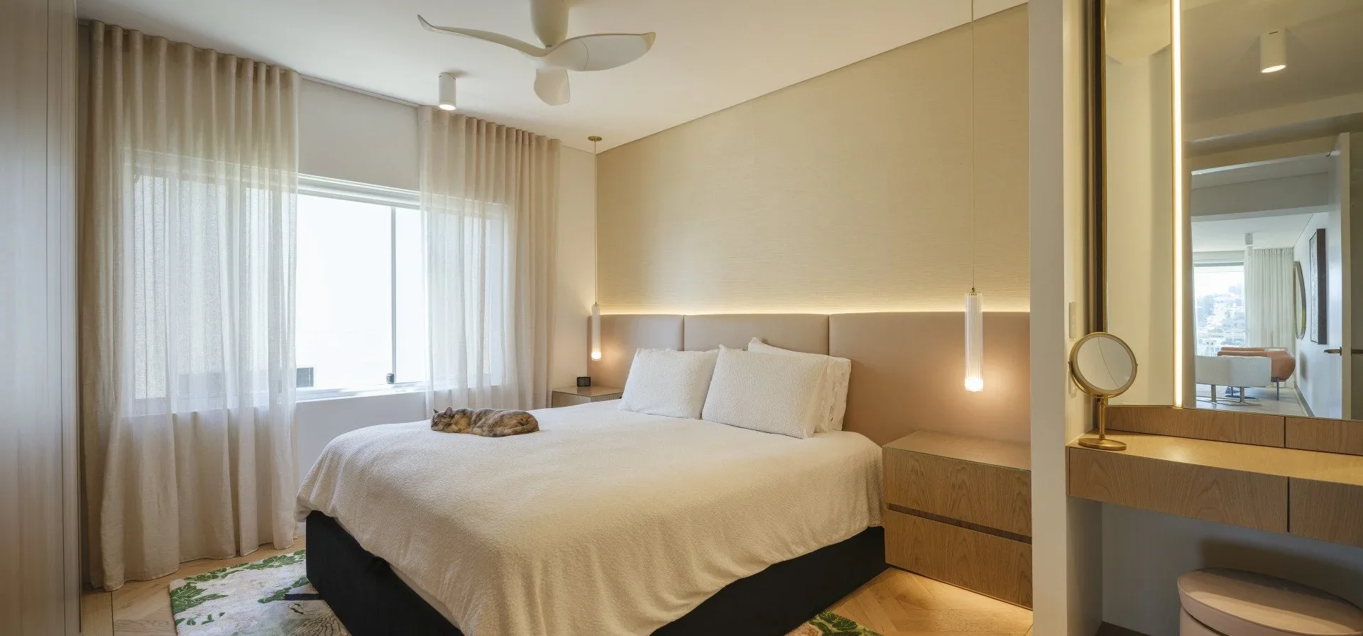 Modern bedroom with a large bed, a cat sleeping on it, light-colored curtains, wooden furniture, and a mirror reflecting a sitting area.