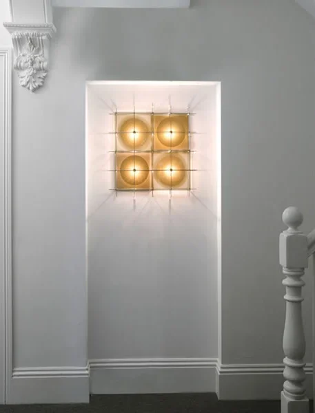 A wall niche with four gold-colored spherical lights arranged in a grid, illuminated inside a white wall with decorative architectural molding on the left and a white staircase railing on the right.