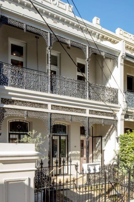 Two-story Victorian-style house with ornate iron balcony and fence, white exterior, and front steps leading to entrance