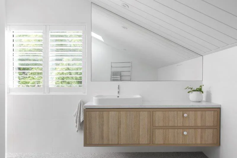 Bathroom with white walls, a large mirror, a white sink, a wooden vanity with drawers, a window with blinds, and a small potted plant.