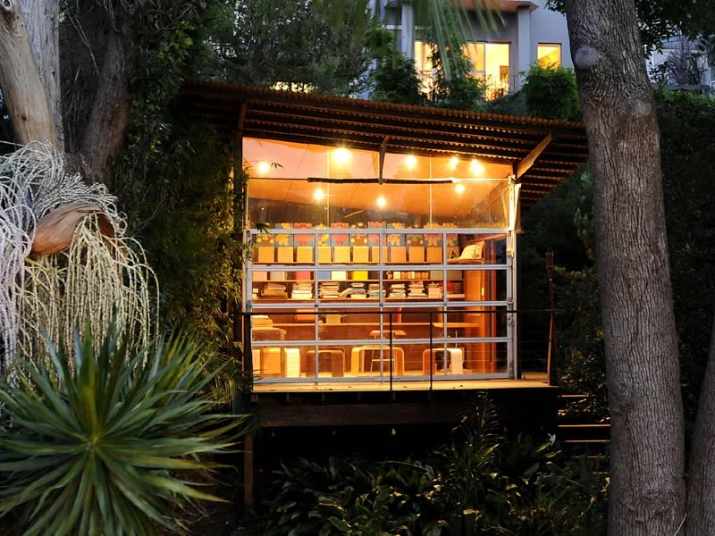A small, modern, glass-walled cafe or workspace surrounded by trees and plants, with warm lighting and a view of books and seating inside.