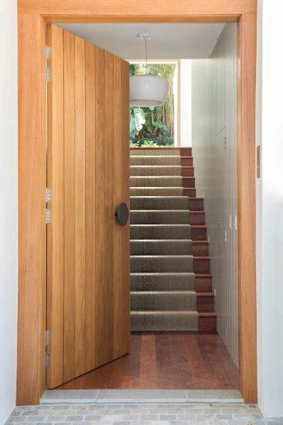 Open wooden door leading to carpeted staircase with a white pot lamp hanging at the top, and greenery visible outside
