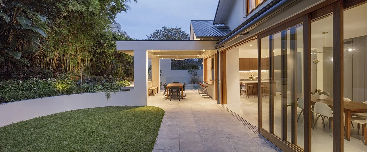 A modern house with an outdoor patio, dining area, and large glass sliding doors revealing an interior with a dining table and chairs.
