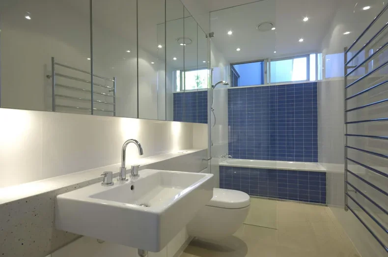 Modern bathroom with a white sink, toilet, and blue-tiled bathtub, large mirror, and heated towel rail, illuminated by ceiling and wall lights.
