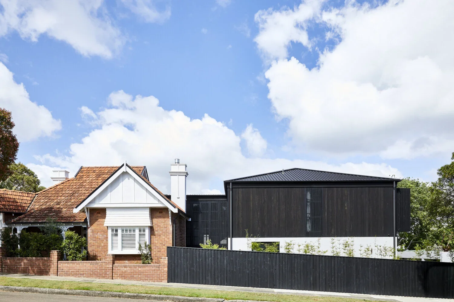 Comparison of traditional brick house with white accents and a modern black house with wooden facade, both behind fences under a blue sky with clouds.