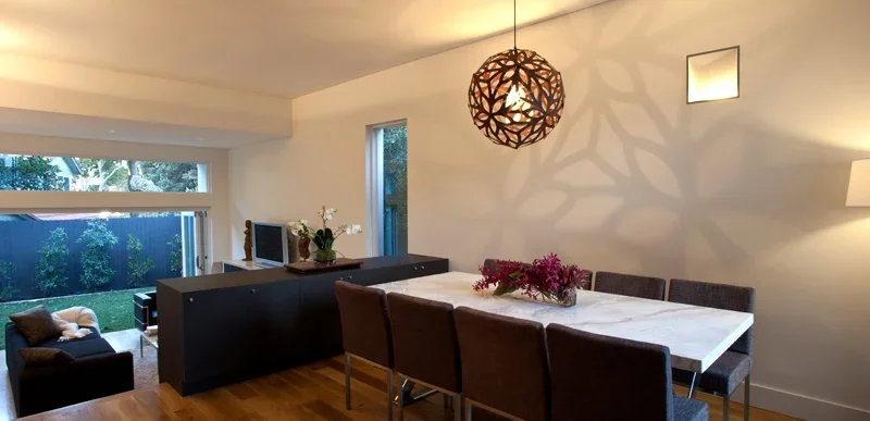 Modern dining room with a white marble table, six dark upholstered chairs, a decorative wooden pendant light, and a small wall-mounted mirror. A black sideboard with a flower arrangement is against the back wall, and large windows overlook a backyard