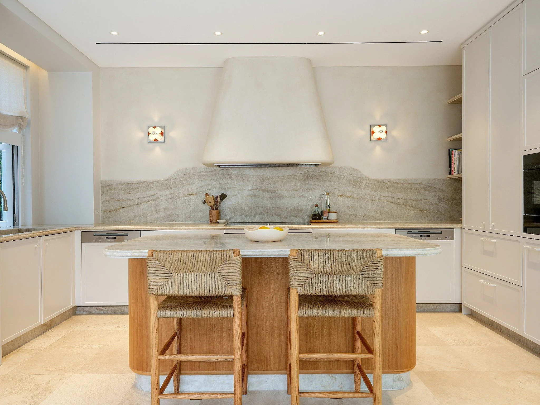 Modern kitchen with white cabinets, marble island, two beige bar stools, grey textured backsplash, and built-in appliances, illuminated by ceiling and wall lights.