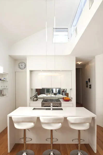 Modern white kitchen with a central island, three white barstools, and a small window above the sink, with natural light coming through a skylight.