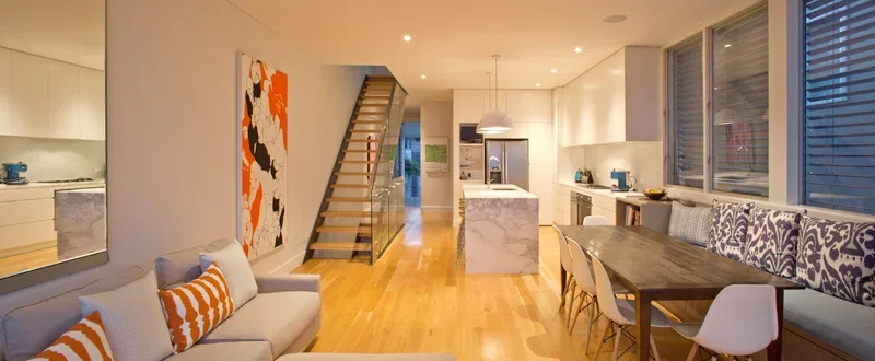 Open-concept living room and kitchen with wooden floors, white cabinetry, marble kitchen island, dining area with wooden table, white chairs, and large windows.