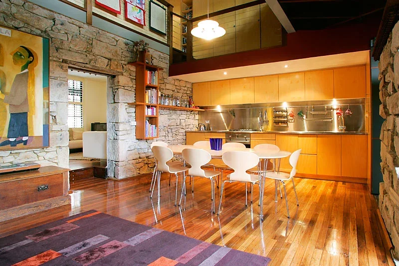 A modern dining area with a wooden table and six white chairs, stone walls, and a kitchen with wooden cabinets and a stainless steel backsplash.