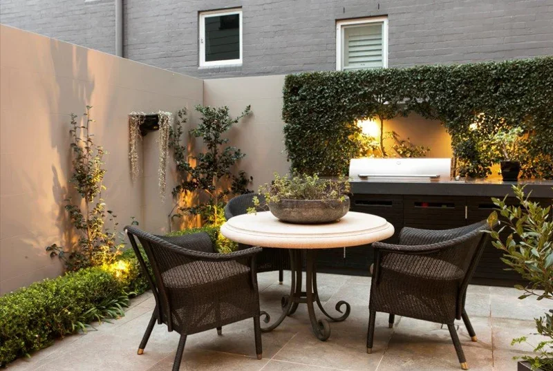 Outdoor patio with a round table, four chairs, garden plants, and a built-in grill under string lights at dusk