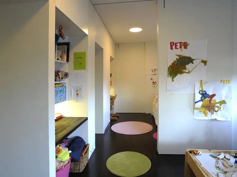 A hallway or play area in a child's room or daycare with children’s artwork on the walls, a pink circular rug, and shelves with toys and books.