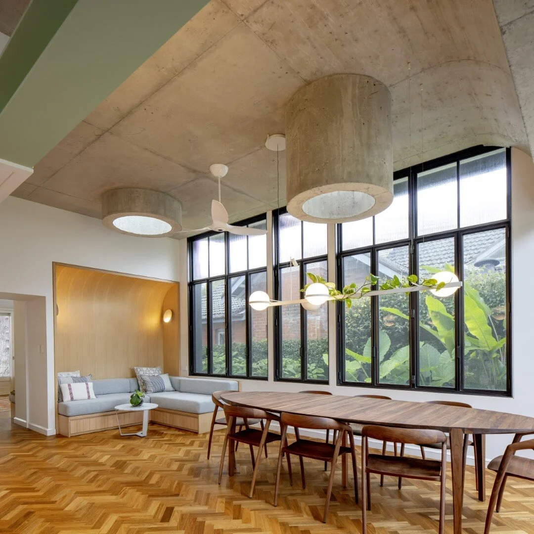 A modern dining room with large black-framed windows, a wooden dining table with matching chairs, a built-in light gray sofa with pillows, and decorative pendant lights with greenery. The ceiling has exposed concrete features, and the floor is patter
