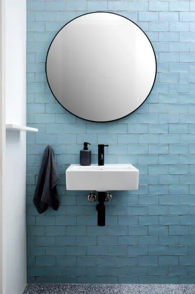 A bathroom wall with a round mirror and a small white sink below it. The wall is covered with light blue bricks. There are black soap dispensers on the sink and a dark towel hanging on a hook to the left.