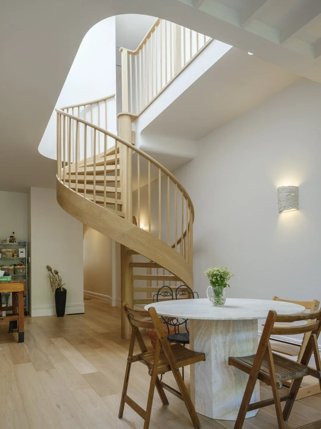 Interior of a home with a wooden spiral staircase, a round dining table with four chairs, a vase with flowers, and a wall sconce light.