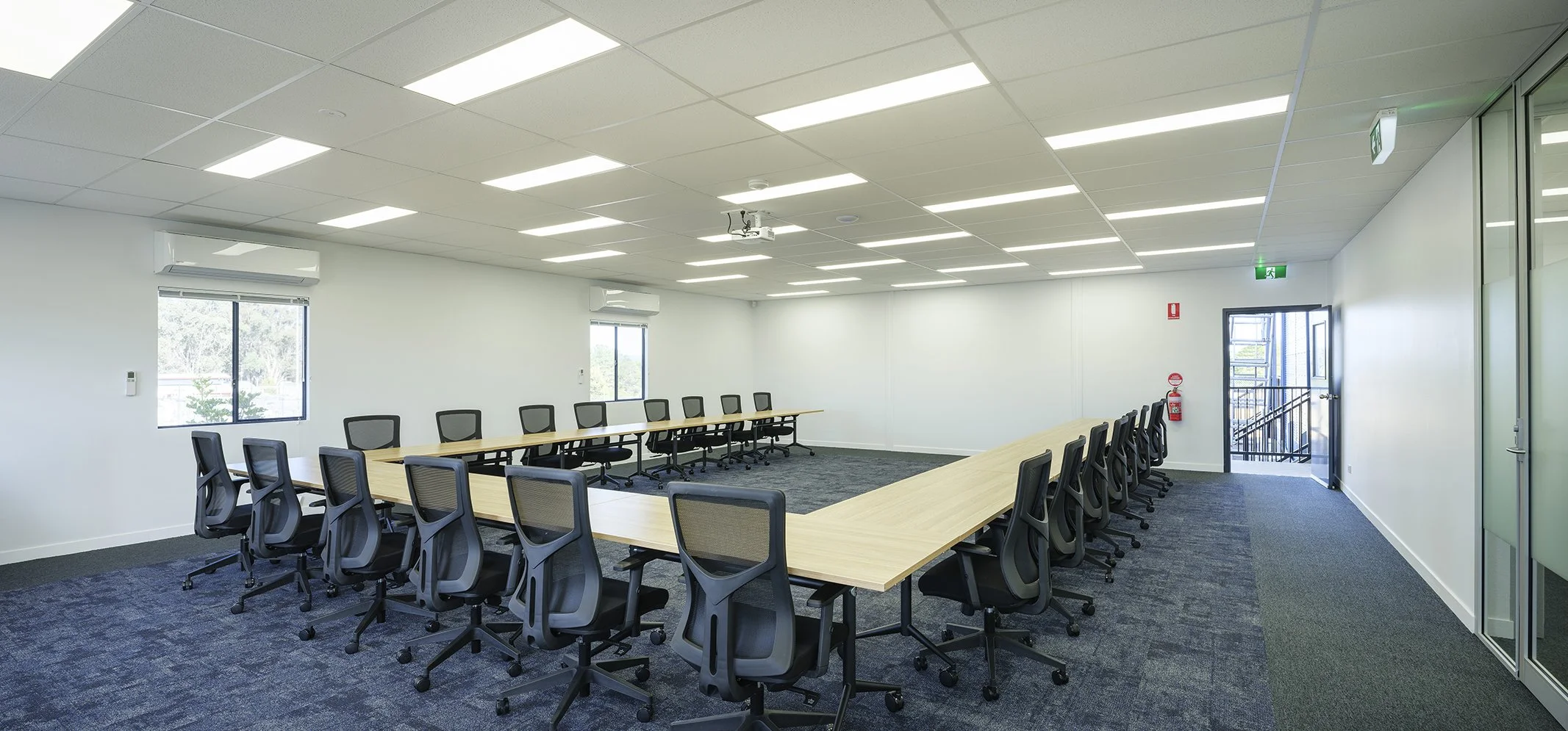 Empty conference room with U-shaped wooden table and black ergonomic chairs, white walls, windows, ceiling lights, and an exit door with emergency signage.