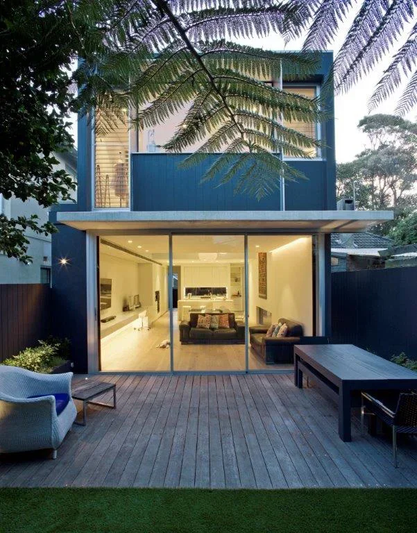 Modern two-story house with glass sliding doors leading to a backyard patio, outdoor furniture including a chair and bench, and trees in the background.