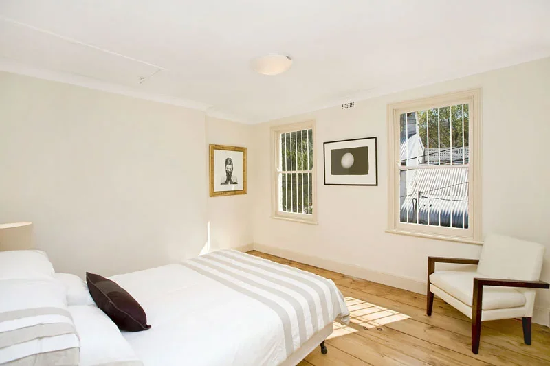 Bright bedroom with white walls, wooden floor, two windows, a white bed with striped bedding and a dark pillow, framed artwork on the wall, and a white armchair.