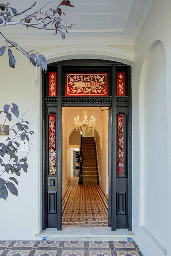 View of an elegant entryway with a partly open decorative black and red door leading into a house, featuring a chandelier hanging and a staircase in the background.