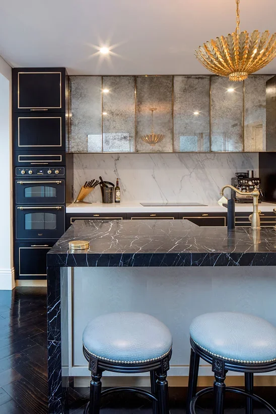 Modern kitchen with black marble island, gold accents, and gray cabinetry, featuring a double oven, bar stools, and a chandelier.