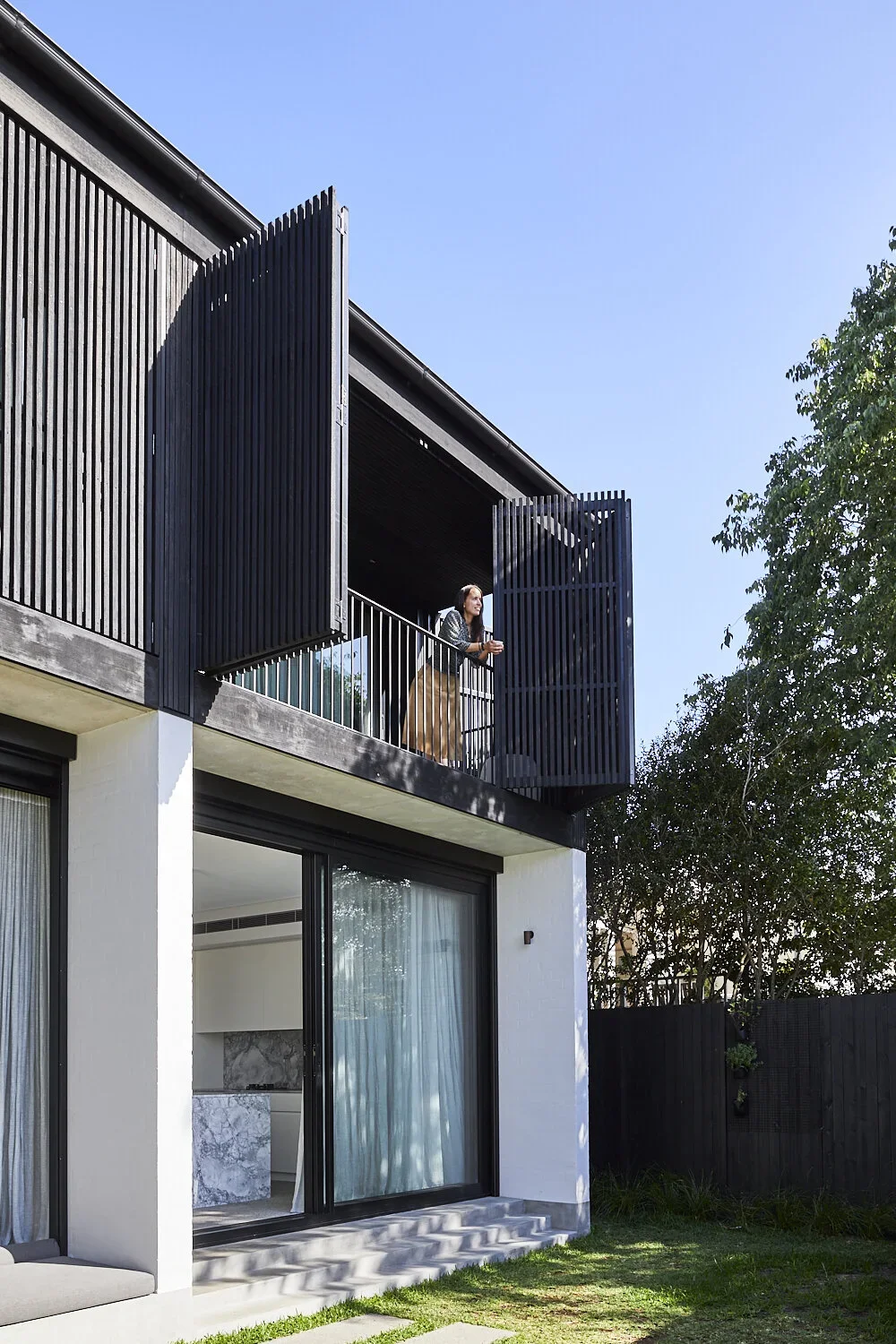 Woman standing on a balcony with modern black wooden slat privacy screens, looking outside on a sunny day.