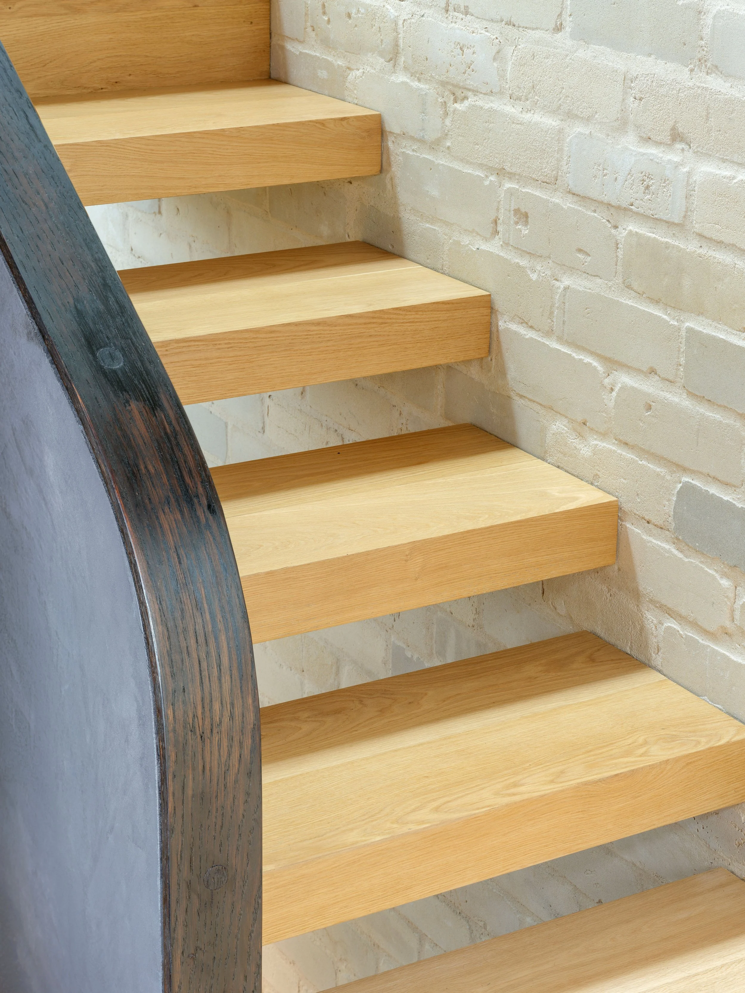 Close-up of pale timber stair treads and a curved handrail beside a light brick wall.