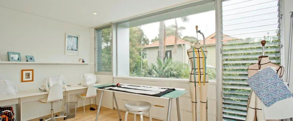 A bright, airy sewing or design studio with a large window overlooking trees and neighboring houses, featuring dress forms, a table for fabric or pattern work, and a white desk with chairs.