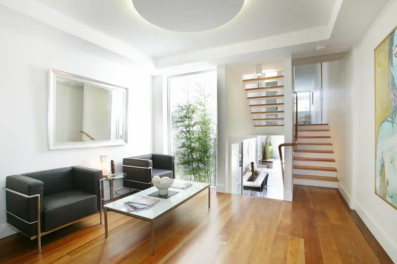 Modern living room with black sofas, glass coffee table, large window with greenery outside, staircase with wooden steps, hardwood flooring, and artwork on the wall.