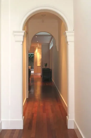 A hallway with wooden flooring and white walls, arched doorway, and a small table with a plant at the end.
