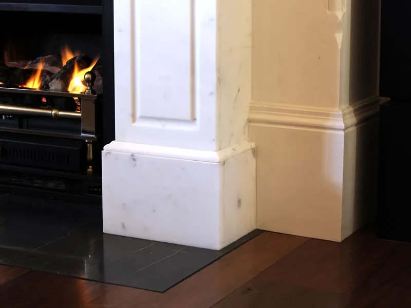 Close-up of a white marble fireplace with a fire burning inside, situated on a dark tiled hearth and adjacent to a wooden floor.